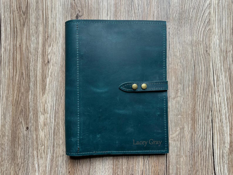 May include: A teal leather journal with a strap closure and brass-colored studs. The journal has stitched edges and the name "Lacey Gray" is embossed on the bottom right corner. The journal is on a wooden surface.