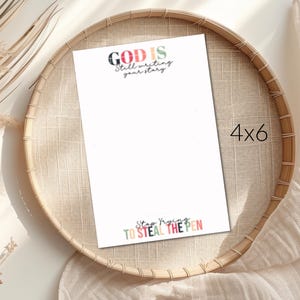 May include: A white 4x6 inch print with the text "GOD IS Still writing your story" at the top and "Stop Trying TO STEAL THE PEN" at the bottom. The print is on a woven tray with dried plants and a beige cloth in the background.