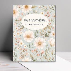 May include: A rectangular greeting card featuring a light sage green background with a pattern of pale pink and white flowers, buds, and green leaves. A central oval contains the black text Love never fails. 1 CORINTHIANS 13:8.