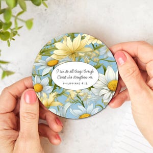 May include: Round coaster with a floral design featuring white daisies and yellow flowers on a light blue background. The coaster has a white oval with the text "I can do all things through Christ who strengthens me. Philippians 4:13"
