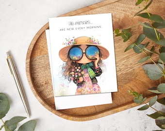 May include: Greeting card with an illustration of a girl wearing a hat and sunglasses, holding flowers. The card has the text "His mercies... are new every morning." The card is on a wooden tray with a pen.
