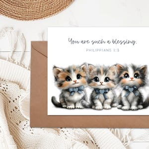 May include: Greeting card featuring three adorable kittens with blue bow ties. The card reads "You are such a blessing. Philippians 1:3". The card is paired with a brown envelope, set against a neutral background.