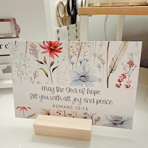 May include: A floral print card with the text "May the God of hope fill you with all joy and peace. Romans 15:13." The card features red, blue, and white flowers and is held by a small wooden block.