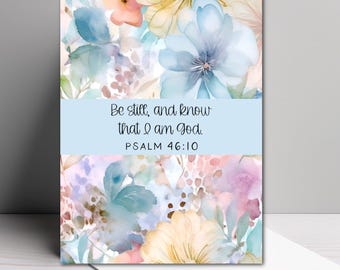May include: A rectangular greeting card featuring a watercolor floral pattern in shades of blue, yellow, pink, and green. A horizontal light blue band across the center displays the black text: Be still, and know that I am God. PSALM 46:10.