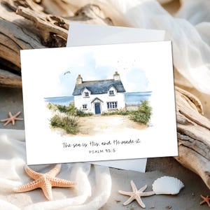 May include: A watercolor print of a white cottage with a blue door and roof, set on a sandy beach with the ocean in the background. The text "The sea is His, and He made it. Psalm 95:5" is printed below the image. Starfish and a seashell are in the foreground.