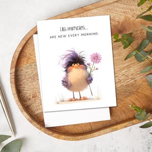 May include: A greeting card featuring an illustrated bird with purple and beige feathers, holding a pink flower. The card has the text "His mercies... are new every morning." The card is on a wooden tray with a pen and greenery.