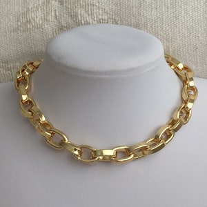 以下が含まれることがあります： 大きな連結リンクが付いたゴールドカラーのチェーンネックレス。 ネックレスは、ニュートラルな背景に対して白いマネキンのバストに表示されています。 チェーンリンクは、研磨された反射面を持っています。