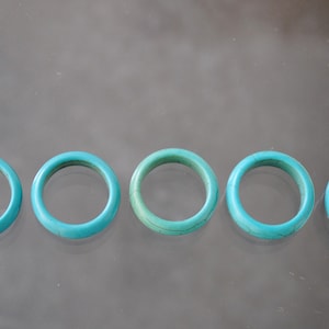 May include: Five turquoise rings, each with a smooth, rounded surface, are arranged in a row on a reflective surface.
