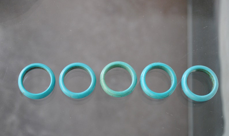 May include: Five turquoise rings, each with a smooth, rounded surface, are arranged in a row on a reflective surface.