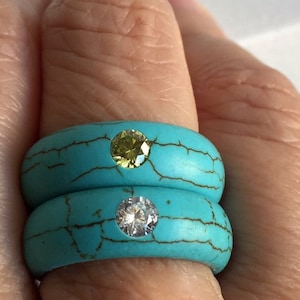 May include: Two turquoise rings with a yellow and a clear stone set in the turquoise. The rings are stacked on top of each other.
