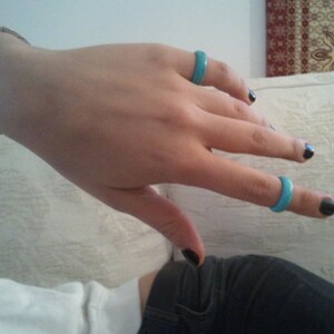May include: A hand wearing three turquoise rings on the fingers. The hand is resting on a white surface. The nails are painted black.