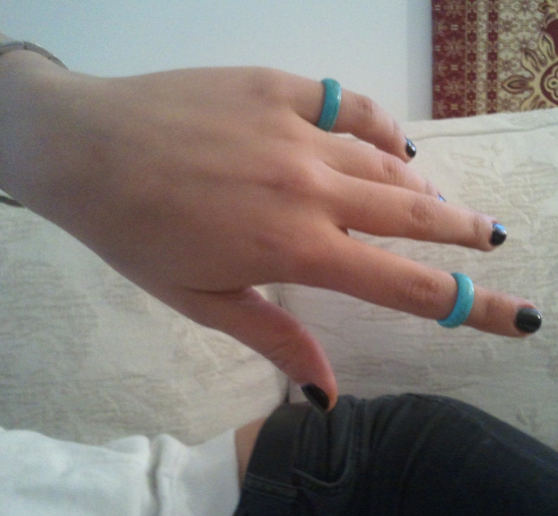 May include: A hand wearing three turquoise rings on the fingers. The hand is resting on a white surface. The nails are painted black.
