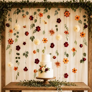 May include: A three-tiered white cake on a wooden pedestal sits on a rustic wooden table. Behind the table, a wooden frame holds hanging strings of orange, burgundy, and cream-colored flowers and green leaves against a white fabric backdrop.