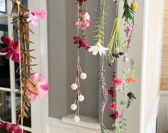 May include: A hanging floral mobile featuring multiple vertical strands of twine adorned with various artificial flowers and greenery, including pink, red, white, and orange blossoms, along with small white spherical beads interspersed throughout.