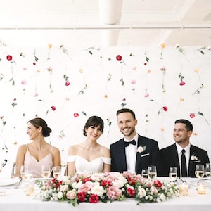 May include: A wedding reception table is set with a white tablecloth, glassware, and floral centerpieces. The wedding party is seated around the table. The backdrop features a white wall adorned with hanging flowers. The bride and groom are in the center.