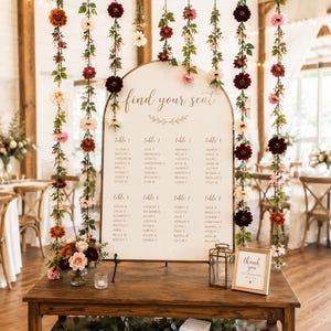 May include: A wooden table displays an arched seating chart sign with the text find your seat and guest names for tables 1 through 8. Hanging floral vines in shades of red, pink, and white drape from a branch above. A small thank you sign sits nearby.