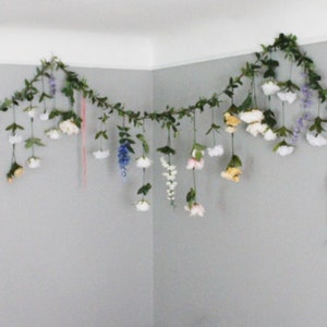 Pu&ograve; includere: Una ghirlanda decorativa di finte foglie e fiori, appesa in un angolo. La ghirlanda presenta una variet&agrave; di fiori bianchi, rosa, gialli e viola, con elementi pendenti. Le foglie sono di un verde intenso, creando un'estetica naturale.