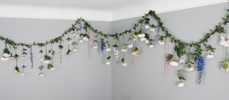 Pu&ograve; includere: Una ghirlanda decorativa di finte foglie e fiori, appesa in un angolo. La ghirlanda presenta una variet&agrave; di fiori bianchi, rosa, gialli e viola, con elementi pendenti. Le foglie sono di un verde intenso, creando un'estetica naturale.