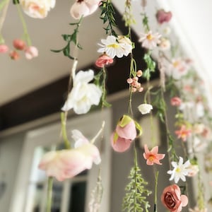May include: A hanging floral installation featuring a variety of artificial flowers in shades of pink, white, and peach, suspended on thin strings. The arrangement includes daisies, roses, and small buds with green foliage, creating a cascading effect.