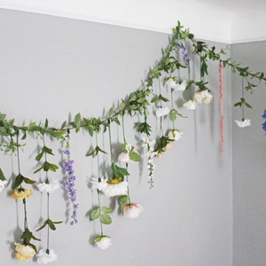 Pu&ograve; includere: Una ghirlanda di fiori e verde artificiale &egrave; appesa a una parete grigia. La ghirlanda presenta fiori bianchi, gialli, rosa e blu, con foglie e viti verdi. Un elemento decorativo.