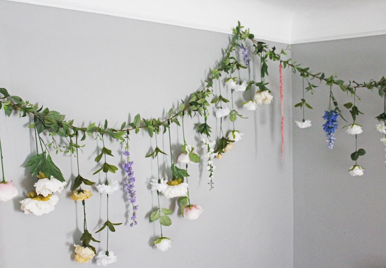 Pu&ograve; includere: Una ghirlanda di fiori e verde artificiale &egrave; appesa a una parete grigia. La ghirlanda presenta fiori bianchi, gialli, rosa e blu, con foglie e viti verdi. Un elemento decorativo.