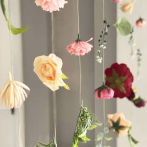 May include: A hanging floral decoration featuring various artificial flowers and green leaves suspended on thin strings. The arrangement includes pink, cream, and deep red blossoms, along with delicate fern-like foliage and small green sprigs.