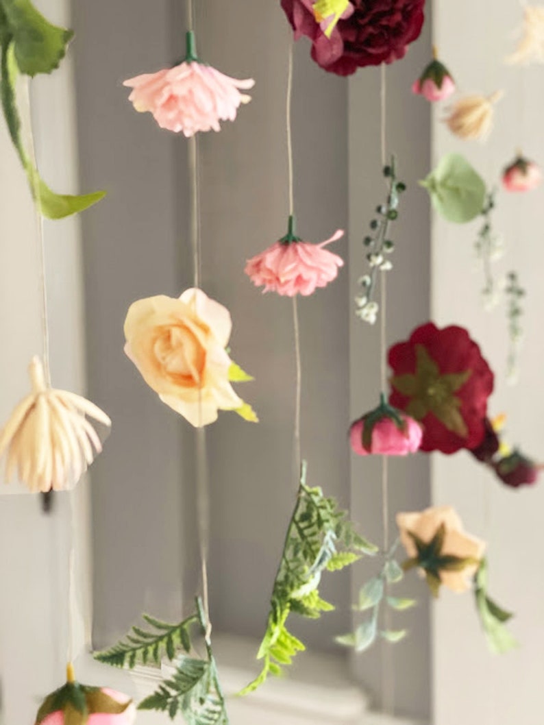 May include: A hanging floral decoration featuring various artificial flowers and green leaves suspended on thin strings. The arrangement includes pink, cream, and deep red blossoms, along with delicate fern-like foliage and small green sprigs.