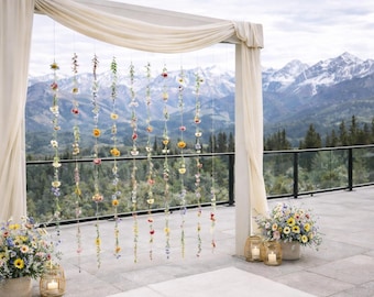 May include: An outdoor wedding ceremony setup with a mountain backdrop. A draped archway is adorned with hanging floral arrangements. Floral bouquets and lanterns with candles flank the arch. The scene is set on a stone patio with a glass railing.