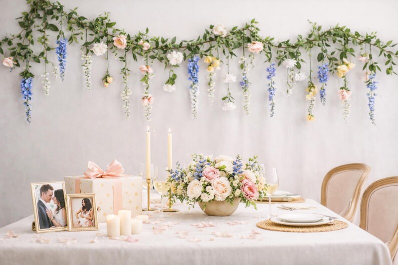 Pu&ograve; includere: Una tavola splendidamente decorata con un centrotavola floreale, candele e foto incorniciate. Sopra, una ghirlanda di fiori e verde pende contro un muro bianco. La tavola &egrave; apparecchiata per un'occasione speciale.