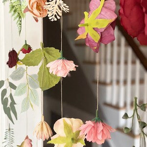 May include: A hanging floral mobile featuring various artificial flowers and foliage suspended on thin strings. The arrangement includes peach, pink, red, and cream-colored blossoms, along with green leaves and fern-like sprigs, creating a cascading display.