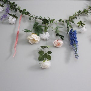 Pu&ograve; includere: Una ghirlanda floreale decorativa con vari fiori e verde artificiali. La ghirlanda comprende fiori bianchi, rosa e blu, insieme a foglie verdi. Una catena di perline rosa &egrave; appesa.