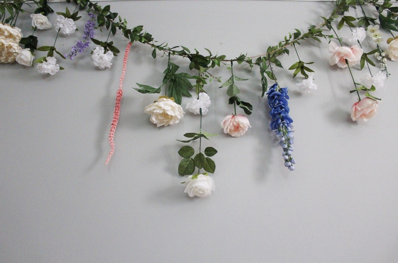 Pu&ograve; includere: Una ghirlanda floreale decorativa con vari fiori e verde artificiali. La ghirlanda comprende fiori bianchi, rosa e blu, insieme a foglie verdi. Una catena di perline rosa &egrave; appesa.