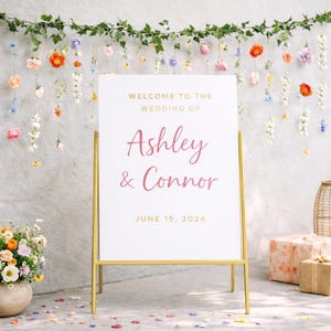 May include: A wedding welcome sign with gold frame, displaying "Welcome to the Wedding of Ashley & Connor, June 15, 2024." The sign is set against a gray wall decorated with floral garlands and hanging flowers. A floral arrangement and gifts are in the foreground.