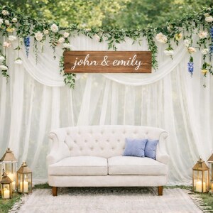 Pu&ograve; includere: Sfondo per cerimonia nuziale con un divano bianco trapuntato e due cuscini blu, su una tenda bianca trasparente. Un cartello di legno recita "john & emily". Lo sfondo &egrave; decorato con fiori, verde e lanterne illuminate.