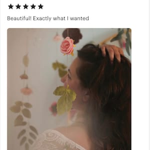 May include: A vertical string of artificial flowers featuring a soft pink rose at the top, followed by green leaves and additional small floral elements suspended on a thin cord. The arrangement is designed to hang vertically as a decorative piece.