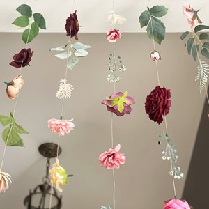 May include: A hanging floral garland featuring multiple vertical strings adorned with artificial flowers in shades of burgundy, dusty pink, and cream, interspersed with green leaves and small white sprigs, suspended from a horizontal line.
