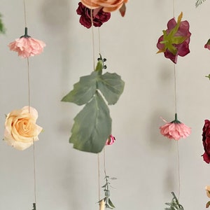 May include: A hanging floral garland featuring various artificial flowers in shades of peach, pink, and deep red, interspersed with green fern leaves and foliage. The flowers and leaves are attached to thin, vertical strings, creating a cascading display.