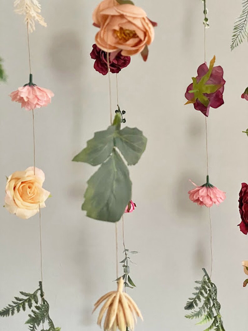 May include: A hanging floral garland featuring various artificial flowers in shades of peach, pink, and deep red, interspersed with green fern leaves and foliage. The flowers and leaves are attached to thin, vertical strings, creating a cascading display.