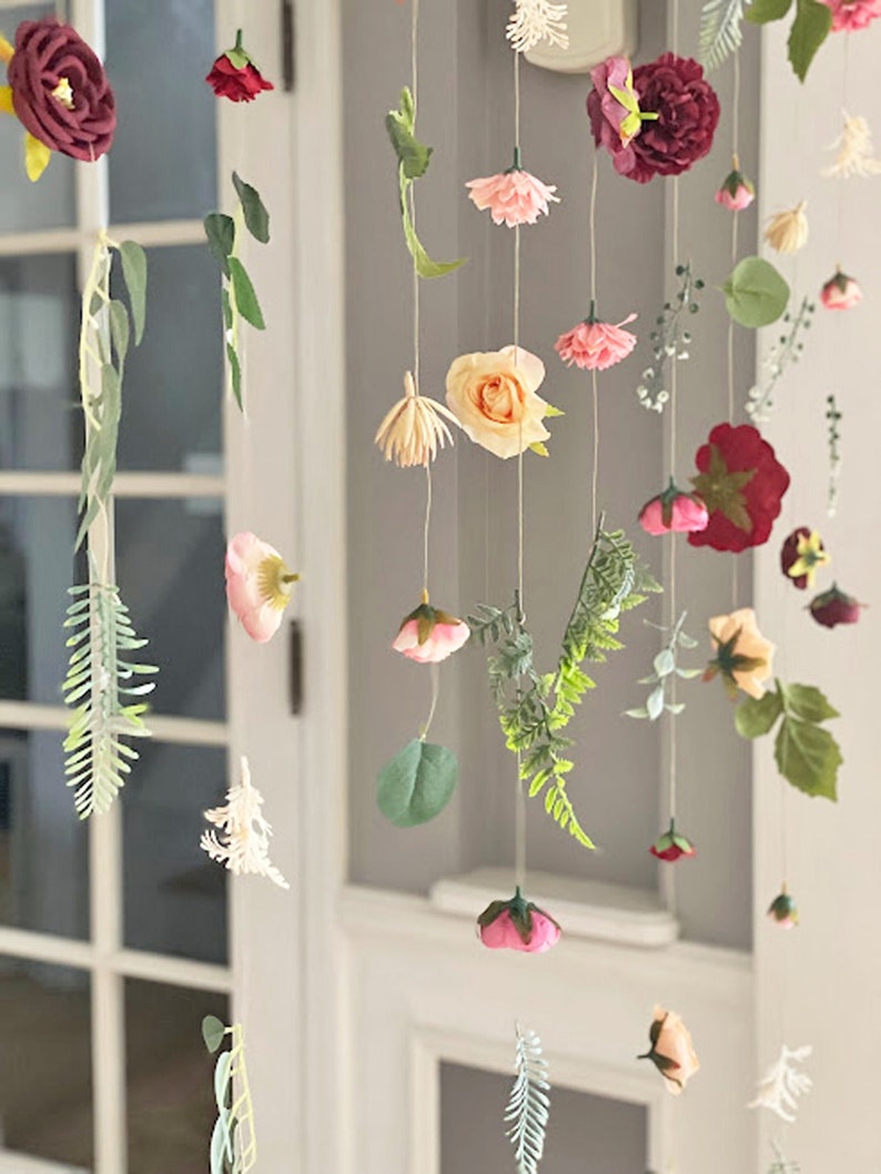 May include: A hanging floral mobile featuring various artificial flowers and green leaves suspended on thin strings. The arrangement includes deep red, light pink, and peach-colored blossoms, along with delicate fern-like foliage and greenery.