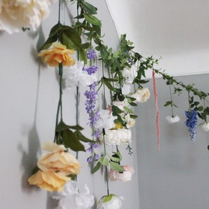 Pu&ograve; includere: Una ghirlanda floreale decorativa con una variet&agrave; di fiori artificiali, tra cui rose bianche, gialle e rosa, e glicini viola. Foglie e viti verdi sono sparse nell'arrangiamento, creando un'estetica naturale e decorativa.
