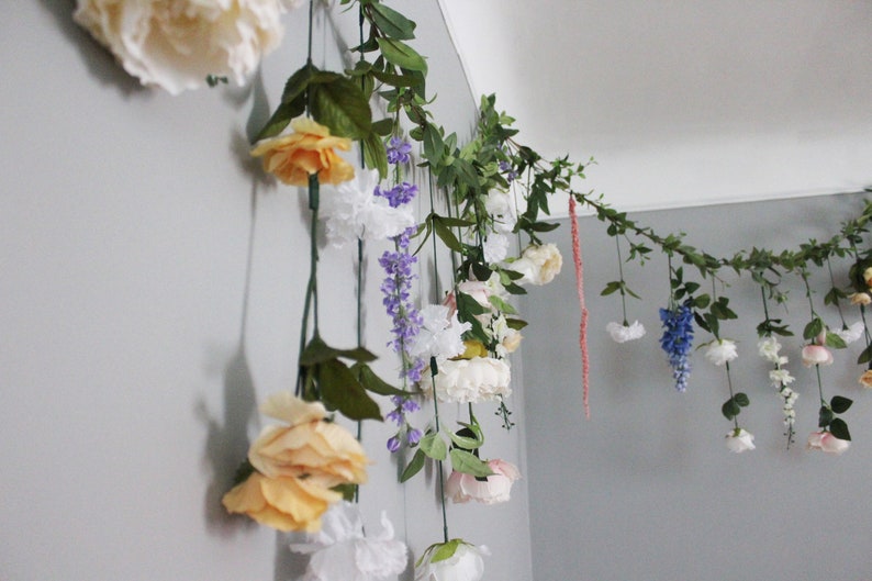 Pu&ograve; includere: Una ghirlanda floreale decorativa con una variet&agrave; di fiori artificiali, tra cui rose bianche, gialle e rosa, e glicini viola. Foglie e viti verdi sono sparse nell'arrangiamento, creando un'estetica naturale e decorativa.