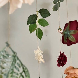 May include: A hanging floral mobile featuring artificial roses in shades of deep burgundy, soft peach, and light pink. The arrangement includes green foliage, small white sprigs, and delicate fern leaves suspended on thin strings, creating a layered display.