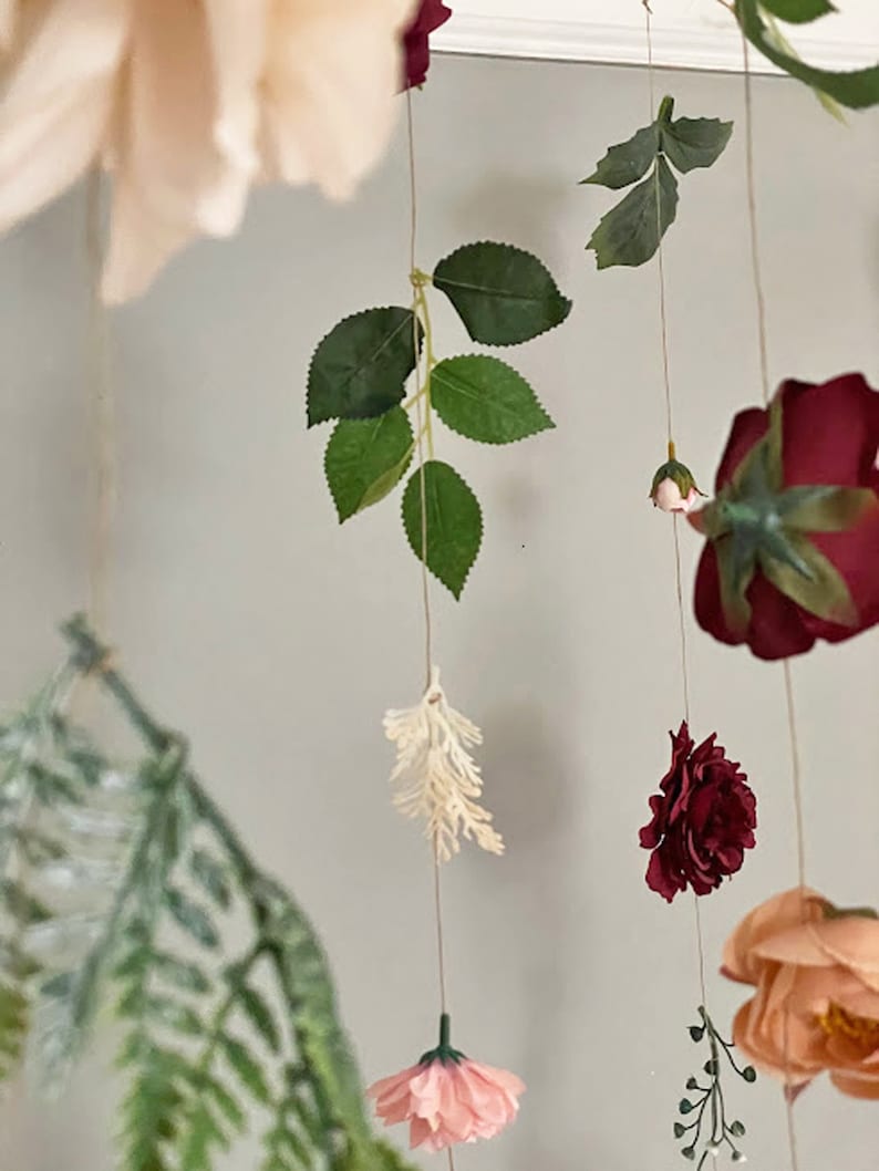 May include: A hanging floral mobile featuring artificial roses in shades of deep burgundy, soft peach, and light pink. The arrangement includes green foliage, small white sprigs, and delicate fern leaves suspended on thin strings, creating a layered display.
