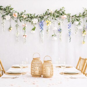 Pu&ograve; includere: Un tavolo da pranzo apparecchiato per un'occasione speciale, con una tovaglia bianca, lanterne intrecciate e sedie in bamb&ugrave;. Una ghirlanda floreale con fiori dai colori pastello &egrave; appesa sopra il tavolo contro un muro bianco. Il tavolo &egrave; apparecchiato con piatti, bicchieri e posate.