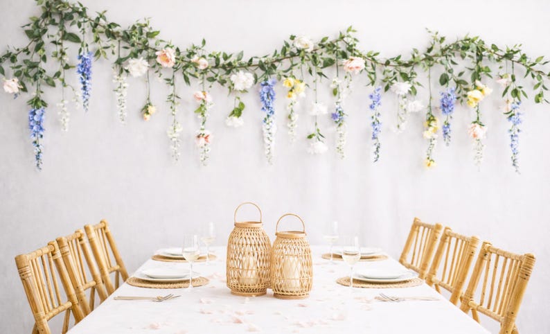 Pu&ograve; includere: Un tavolo da pranzo apparecchiato per un'occasione speciale, con una tovaglia bianca, lanterne intrecciate e sedie in bamb&ugrave;. Una ghirlanda floreale con fiori dai colori pastello &egrave; appesa sopra il tavolo contro un muro bianco. Il tavolo &egrave; apparecchiato con piatti, bicchieri e posate.