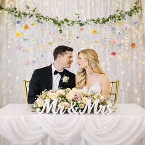 May include: A wedding reception scene with a couple seated at a table. The table is decorated with a floral arrangement and a "Mr & Mrs" sign. The background features a curtain with fairy lights and floral decorations.