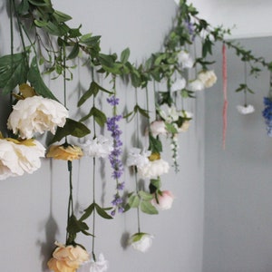 Pu&ograve; includere: Una ghirlanda floreale con fiori appesi nei toni del bianco, rosa, giallo e viola. La ghirlanda &egrave; composta da fiori e verde artificiali, creando un'esposizione decorativa. I fiori sono attaccati a un tralcio.