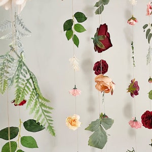 May include: A hanging floral garland featuring multiple vertical strings adorned with artificial roses in shades of deep red, peach, and soft pink, interspersed with green fern leaves and small white sprigs, all suspended on thin, light-colored twine.