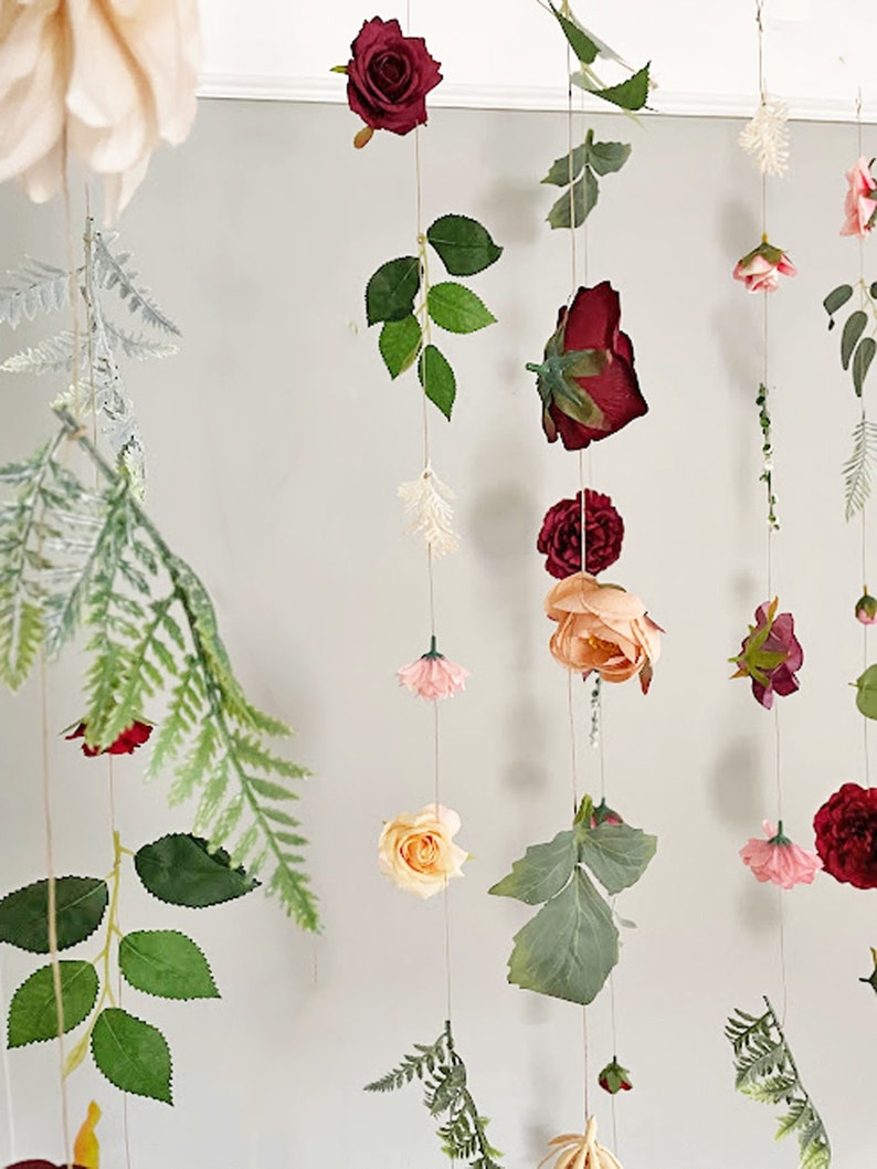 May include: A hanging floral garland featuring multiple vertical strings adorned with artificial roses in shades of deep red, peach, and soft pink, interspersed with green fern leaves and small white sprigs, all suspended on thin, light-colored twine.