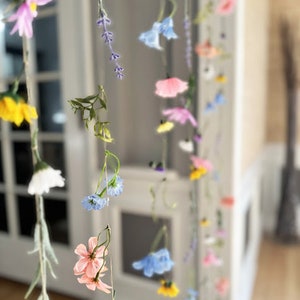 May include: A hanging floral garland featuring multiple vertical strands of artificial flowers in various colors including pink, yellow, blue, white, and purple. The delicate blossoms and green foliage are suspended on thin strings, creating a cascading effect.